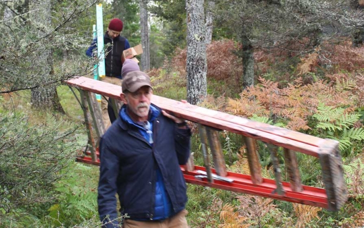 Two people carrying ladders walk through a wooded area during a service project with Outward Bound.